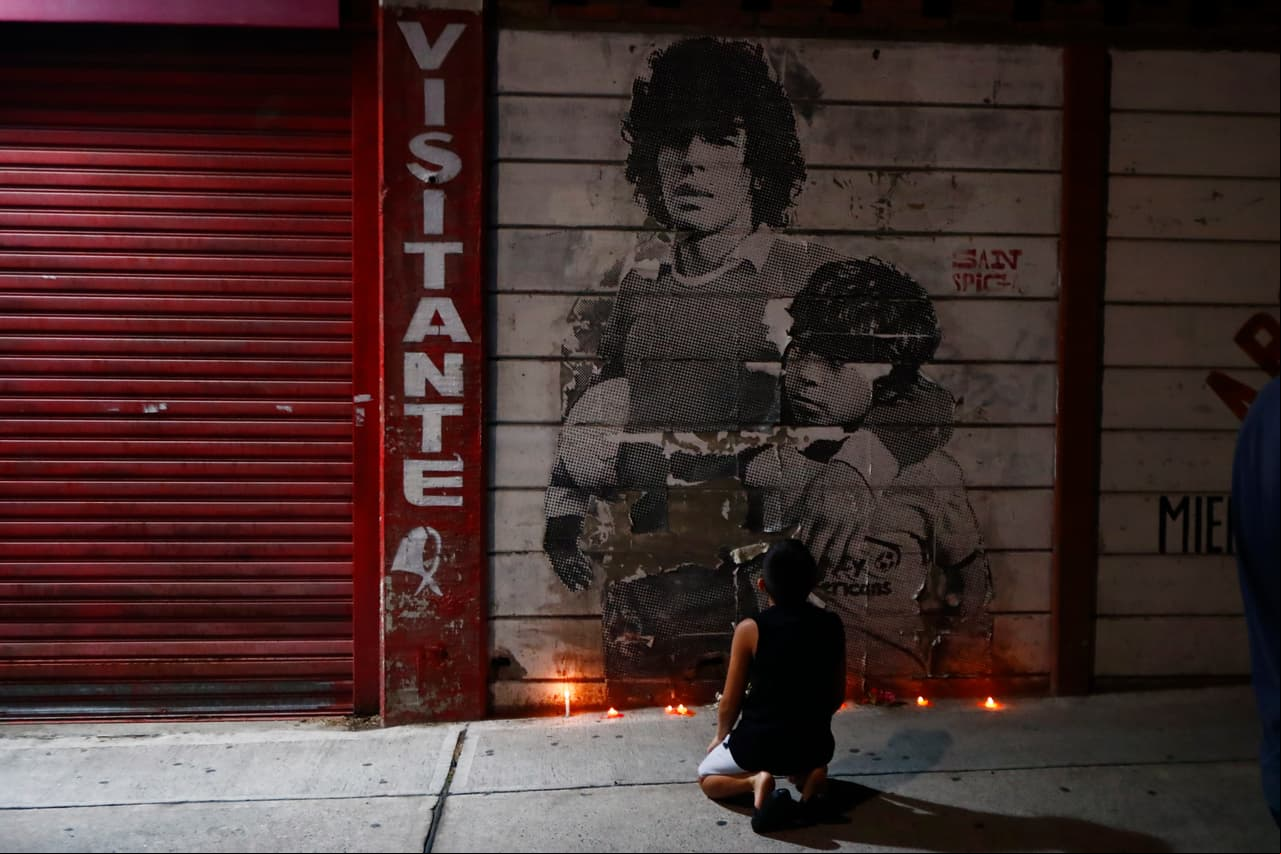 Un niño se arrodila frente a un mural en el que aparece Maradona en sus primeros años como futbolista abrazando a uno de sus hermanos.